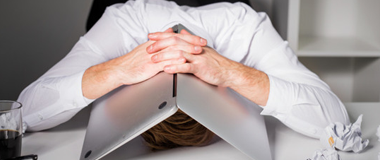 A man with his face pressing against the table looking stressed, with his laptop folded over his head